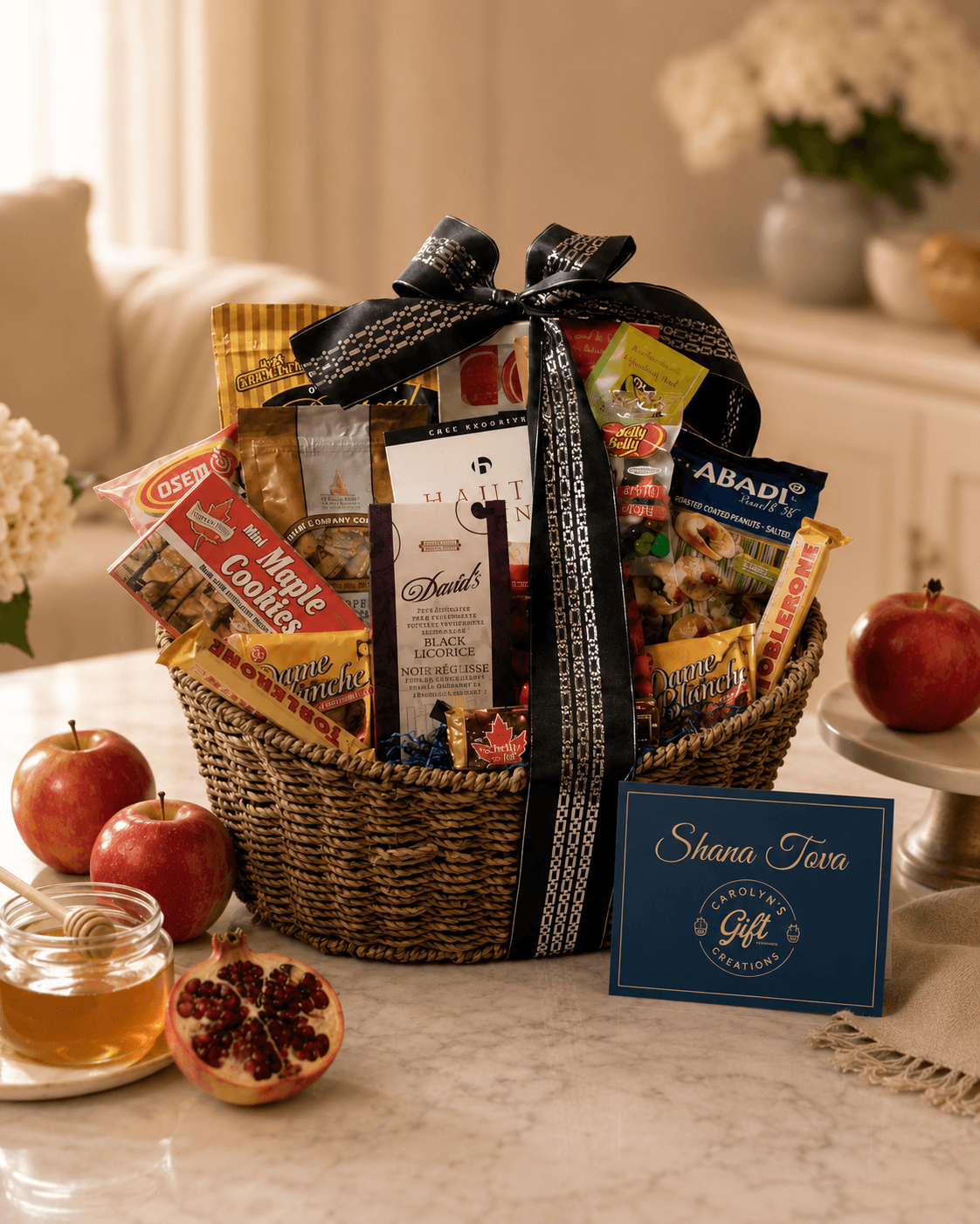 A woven gift basket filled with assorted snacks, chocolate, and packaged treats, tied with a black ribbon. The basket sits on a marble table with apples, honey, and a blue gift card reading 'Shana Tree'.