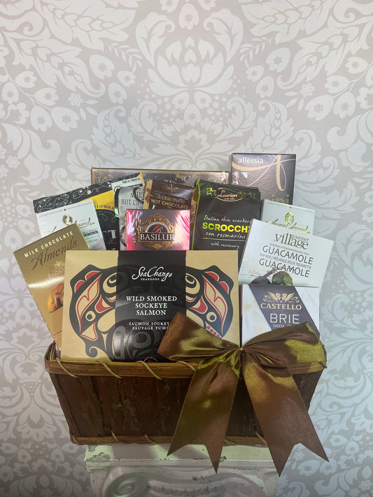 A gourmet chocolate gift basket filled with assorted packaged chocolates and sweets, tied with a large metallic gold ribbon, set against a light gray patterned fabric background.