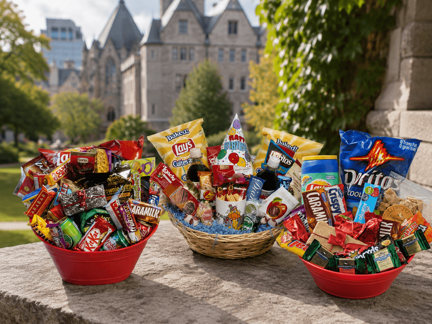 Three snack gift baskets filled with assorted packaged foods, placed on a stone ledge outdoors with a large historic building and greenery in the background. Visible snack brands include Dritzos, Lay's, KitKat, Armstrong, and Ritz.