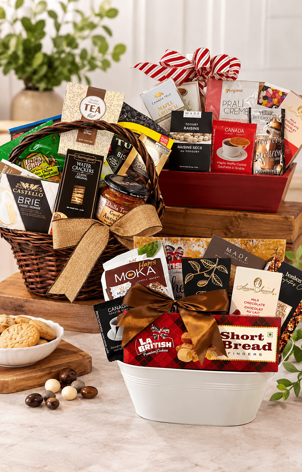 A gourmet gift basket arrangement featuring three baskets filled with assorted packaged snacks, teas, coffee, and breads, styled on a marble surface with a bowl of cookies and greenery in the background.